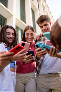 Dikey. Dışarıdaki cep telefonundan gülümseyen videolara bakan bir grup beyaz gencin alçak açılı görüntüsü. Üç kuşak Z öğrencisi arkadaşlar kampüste birlikte cep telefonu izleyerek eğleniyorlar.