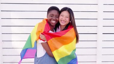 Genç lezbiyen çift, eşcinsel gurur bayrağıyla sarılıyor. Beyaz arka planda kucaklaşan gülümseyen kameraya bakan iki sevimli arkadaş. Asya ve Afrika birleşik homoseksüel aşkı