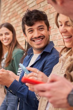 Dikey. Dışarıdaki telefonları kullanarak duvara yaslanan çok ırklı gülümseyen genç nesil Z 'nin yan görüntüsü. Arkadaşlar birlikte sosyal medyaya bakarak eğleniyorlar. İlişkiler ve yeni teknolojiler