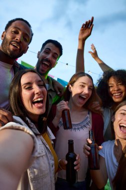 Dikey. Milenyum alkol partisi grubunda selfie çeken neşeli ve çekici genç bir kadın dışarıda kameraya heyecanlı bir şekilde poz veren çok ırklı arkadaşlar. Gece kutlamalarında neşeli insanlar