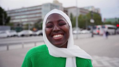 Siyahi Müslüman kadının portresi çok mutlu, gülümseyen ve kameraya bakan. Afrikalı bir öğrenci dışarıda dururken samimi bir şekilde gülümsüyor. Kız beyaz tesettür ve yeşil tişört giyiyor.