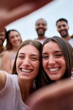 Dikey. İki kız arkadaş ellerle çerçeve yapıyor. Odaklanamayan bir grup genç mutlu bir şekilde selfie çekiyorlar. Gülümseyen kadınlar boş zamanlarında eğleniyorlar.