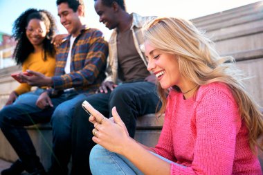 Açık havada cep telefonu kullanan güzel, genç, beyaz, sarışın bir kadın. Bir grup gülümseyen ırklar arası öğrenci akıllı telefona bakıyor. Mutlu milenyum dostları dijital ekranda komik bir şey izliyor.