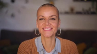 Neşeli, çekici, orta yaşlı, saçları tıraşlı, kameraya gülümseyen bir kadın. Kırklı yaşlardaki neşeli kadınlar evde halka küpeleri ve pozitif yetenekleriyle portre için mutlu poz veriyorlar.