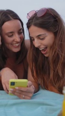 Dikey. İki kız arkadaş plajda sosyal medyayı kontrol etmek için mobil bir cihazı paylaşıyorlar. Genç kadınlar akıllı telefonları kontrol eder ve çok gülerler. Telefonda video izlerken birlikte gülümserler..