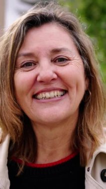 Dikey. Neşeli, olgun, beyaz kadın dışarıda gülümsüyor. Yaşlı sarışın kadın fotoğraf için kameraya mutlu bir poz veriyor.