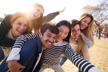Büyük gülücükler ve şakacı hareketlerle bir grup genç parkta eğlenceli bir fotoğraf için poz veriyor, bir ağacın altında boş bir anı paylaşıyorlar.