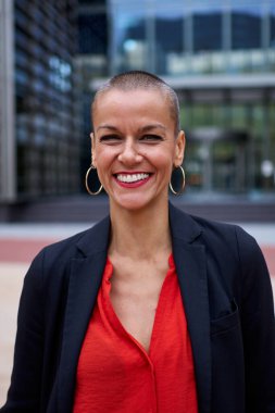 Dikey. Tıraşlı orta yaşlı beyaz takım elbiseli iş kadını modern ofis alanının dışında poz veriyor. Güçlü ve güçlü kırmızı elbiseli çekici kadın iş yerinin önündeki kameraya gülümsüyor.