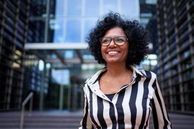 Yetişkin Afro-Amerikalı resmi iş kadınının modern ofis binasının önünde mutlu bir jestle dik dik dik durduğu sahneyi kopyalayın. Profesyonel zarif siyah kadın dışarıda poz veriyor.