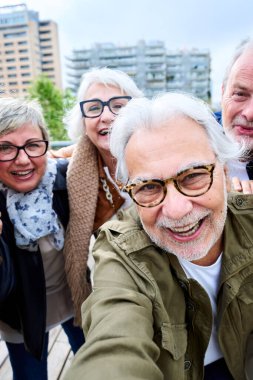 Dikey. Selfie grubu olgun Kafkasyalıların arka planda birlikte gülümseyen fotoğraflar çekmesi. Yaşlı gri saçlı şehir arkadaşları dışarıda kameraya mutlu görünerek hatıra fotoğrafı çekmek için telefon kullanıyorlar.
