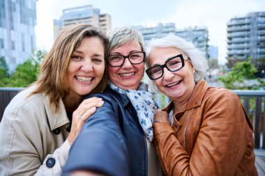 Kentte duran mutlu fotoğraf pozu veren üç olgun kadının selfie portresi. Yaşlı, gri saçlı turistler. Dışarıda tatilde kameraya gülümseyen kadınlar. 