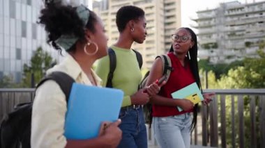 Üniversite kampüsüne giden tek siyahi kız öğrencilerin yan görüntüsü. Üç genç Afrikalı Amerikalı mutlu kadın arkadaş rahatça yürüyüp sırt çantaları ve kitaplarla şehirde sohbet ediyorlar.