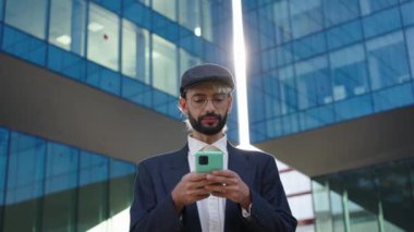 Güneşli bir günde ofisin dışında cep telefonuna bakarak rahatça dolaşan Kafkasyalı genç iş adamının düşük açılı görüntüsü. Profesyonel şehir alternatifi erkek, arka planda bina ve cep telefonu kullanarak mutlu bir şekilde yürüyor