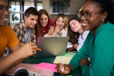 Bir grup genç üniversite öğrencisi, kampüs çalışma salonunda dizüstü bilgisayarla toplandı. Çok ırklı Z jenerasyonundan insanlar ders programını inceliyor. Bir fakülte masasında yan yana sırıtarak oturuyorlar.