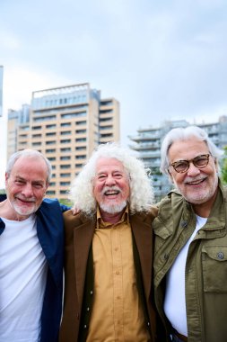 Dikey. Neşeli üç beyaz erkek arkadaşın portresi. Şehirde sarılarak mutlu poz veriyorlar. Yaşlı gri saçlı turistler. İnsanlar tatilde dışarıda kameraya bakıp gülümsüyor. 