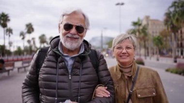 Kamera, beyaz, yaşlı ve aşık bir turist çiftin ellerinde bavul tutarken kameraya gülümserken görüntüsünü büyütür. Gri adam ve kadın birlikte kış turizminin tadını çıkarıyorlar.