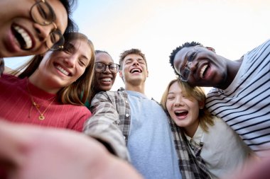 Uluslararası Gen Z arkadaş grubu açık havada güneşli bir gün için heyecanlı görünüyor. Genç öğrenciler gülüyor birbirlerine sarılıyorlar mutlu mesut ayakta durup üniversite tatillerinin tadını çıkarıyorlar. 