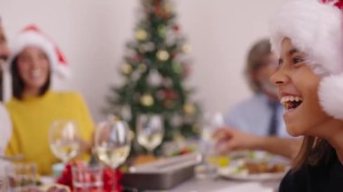 Evde Noel şapkası takmış mutlu aile toplantısına gülen beyaz bir kızın odaklanmış ve odaklanmış olmayan yan görüntüsü. Çeşitli insanlar neşeli bir şekilde masada oturmuş içeride şenlikli bir yemeği kutluyor.