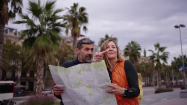 Yetişkin beyaz bir çift ellerinde kağıt harita tutarak mutlu bir şekilde konuşuyor ve turistlerin ilgilendiği yerleri gösteriyorlar. Kadın ve erkek şehir meydanında durmuş turizm tatilinin tadını çıkarırken birlikte gülüyorlar.