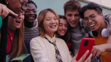 Bir grup mutlu, çok ırklı üniversite öğrencisi açık havada mobil buluşmaya gülüyorlar. Asyalı kızın kırmızı telefonuyla çektiği selfie videosu için poz veren neşeli arkadaşlar. 