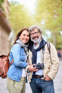 Dikey. Şehirde fotoğraf çektirmek için poz veren mutlu yaşlı bir çift portresi. Tatilde birlikte seyahat eden gülümseyen yaşlılar. Erkek ve kadın, hafta sonu kaçamağında şefkatle sarılıyorlar.