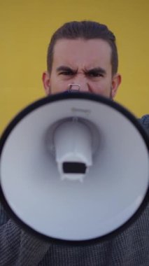 Dikey. Kafkasyalı genç adam izole edilmiş sarı arka planda megafonla öfkeyle bağırıyor. Erkek protestocu sert bir hareketle kameraya bakıyor. İnsanlar gösteri yapıyor. 