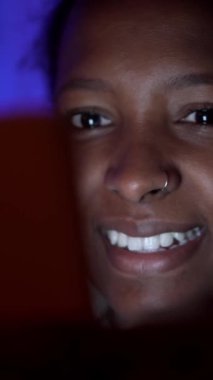 Geceleri cep telefonunu sosyal medya için kullanan güzel Afro-Amerikan kadının dikey yakın çekimi. Neşeli siyah kuşak z kız yüzü gülüyor ağlara bağımlı.