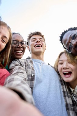 Dikey. Selfie Uluslararası Gen Z arkadaş grubu açık havada güneşli bir günde kamera karşısında heyecanlı görünüyor. Genç öğrenciler gülüyor birbirlerine sarılıyorlar mutlu mesut ayakta durup üniversite tatillerinin tadını çıkarıyorlar. 