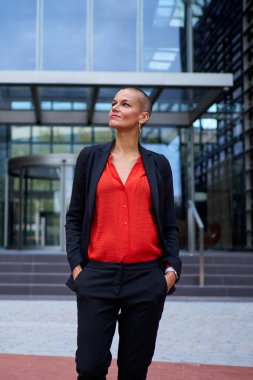 Dikey. Yetişkin resmi iş kadını, modern ofis binasının önünde kendine güvenen bir jestle yukarı bakıyor. Açık havada liderlik yapan çekici, tıraşlı bir kadın.