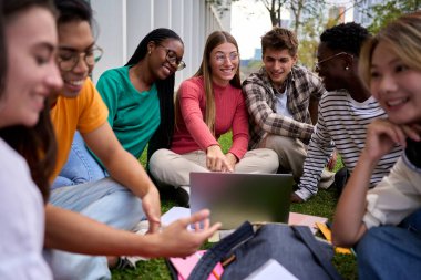 Çeşitli nesiller z Üniversitesi neşeli öğrencileri kampüs parkında çimlerin üzerinde oturan müfredattan geçiyorlar. Arkadaşlar güler yüzlü sınıf arkadaşlarıyla buluşur birlikte ders çalışır ve dışarıda dizüstü bilgisayar kullanır.