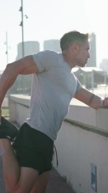 Dikey. Spor giyimli yetişkin beyaz erkek, güneşli havada koşuya çıkmadan önce ısınmak ve ısınmak suretiyle günlük spora hazırlanıyor. Sağlıklı, olgun, yakışıklı bir sporcu tek başına antrenman yapmaktan hoşlanıyor.