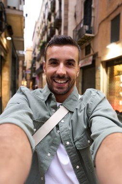 Arka planda Barcelona City turistik caddesinde selfie çeken dikey bir genç adam gülümsüyor. Milenyum erkek turist iki eliyle telefonunu tutarken mutlu bir fotoğraf çekiyor.