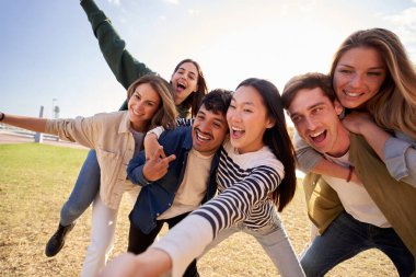 Bir grup genç, bir tarlada mutlu bir anı paylaşıyor, büyük gülücüklerle poz veriyor ve başparmaklarını kaldırıyor, açık mavi gökyüzünün altında, kabarık bulutlarla.