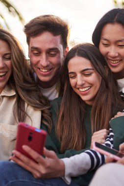 Dikey. Gezici selfie için mutlu poz veren bir grup genç dışarıda oturuyor. Çok ırklı Z kuşağı arkadaşları, güneşli bir günde, tatil fotoğrafı için cep telefonlarına bakıp gülümsüyor.
