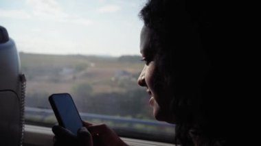 Trenin içinde cep telefonu kullanan ve görünüşlü, gülen Afro-Amerikan bir kadın. Gezerken sosyal ağlarda yazan Z jenerasyonu insanları. Arkaplanda peyzaj alanı