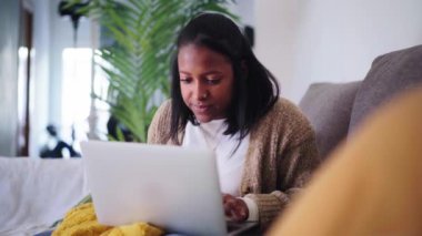 Genç, neşeli Afro-Amerikan kadın dizüstü bilgisayarını kullanıyor evdeki kanepede rahat oturuyor. Oturma odasında sohbet eden, bilgi almak için yazı yazan oldukça siyah bir Z jenerasyonu kadın.