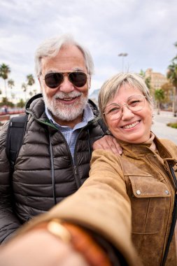 Dikey. Bir çift gülümseyen Kafkasyalı turist birlikte selfie çekiyorlar. Emeklilik tatilinin tadını çıkaran kameraya aşık karı koca. Olgun insanlar turizm yapar.