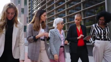 Çeşitli iş kadınlarından oluşan bir ekip ofis binasının yanında tablet kullanarak yürüyor. Resmi giysiler içinde sohbet ediyor ve iletişim kuruyorlar. Bir grup işçi, girişimci ve dışarıda çalışan kadın profesyoneller.