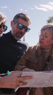 Dikey video. Bir grup bavulu olan turist arkadaşlarıyla orta yaşlı bir çift yürüyor, bakıyor ve bir seyahat haritası tutuyor. Avrupa şehir caddesinde tatil yapan insanların turizmi