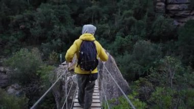 Orman ortamında yemyeşil bir bitki örtüsüyle çevrili ahşap asma köprüsünden yürüyerek geçen tanınmayan kişiyi takip edin. Dağ kıyafetleri içindeki insanlar doğa yürüyüşü yolunda.