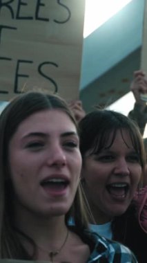 Dikey. Çeşitli gruptan genç sarışın kadın yumrukları havada savaşa karşı öfkeli bağırışları protesto ediyor. Dünya Barış Yürüyüşü gösterilerinde savaş karşıtı pankartlarla yürüyen farklı aktivistler