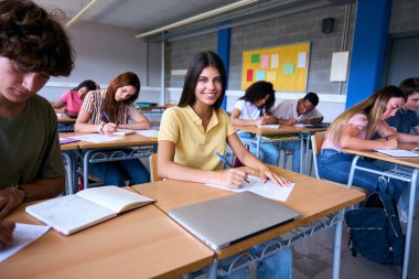 Sınıfta sınıf arkadaşlarıyla birlikte masa başında oturan mutlu beyaz kadın öğrenci portresi. Lisedeki grup arkadaşları derslerine yoğunlaşmışlardı. Ergen bir kadın kameraya bakıp gülümsüyor.