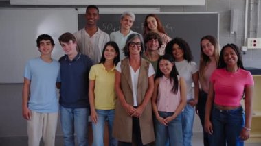 Happy large group of high school student with teacher looking smiling and waving at camera. Young multiracial teenage people posing standing for video in classroom in front of blackboard. Generation z