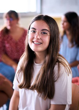 Dikey. Sınıfta arka planda sınıf arkadaşlarıyla oturan Kafkasyalı inek öğrenci portresi. Grup çocukları çalışma odasında toplandı. İlkokulda kameraya mutlu bakan bir kadın pozu.