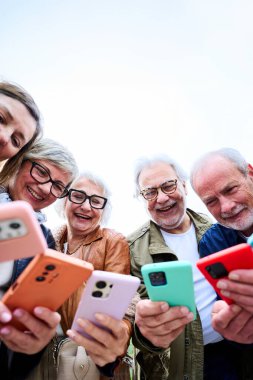 Dikey. Bir grup son sınıf öğrencisi açık havada daireler çizerek renkli akıllı telefonları kullanarak birlikte gülümsüyor. Neşeli olgun gri arkadaşlar gülen cep telefonlarıyla eğleniyorlar. 