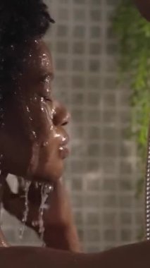 Dikey. Yetişkin afro kadın saçlarını ve başını banyoda duş başlığıyla ıslatıyor. Genç Afro-Amerikalı kadın evinde günlük temizlik için dinleniyor.