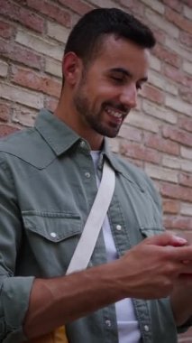 Dikey yakışıklı Avrupalı genç adam cep telefonunu tuğla duvara dayayarak gülümsüyor. Jesti, mutlu yazarken ve dışarı bakarken rahatlamayı öneriyor. Milenyum tarzı erkek