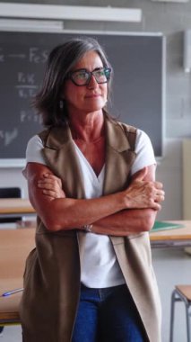 Dikey. Orta yaşlı bayan lise öğretmeni sınıfta kollarını kavuşturup kameraya gülümsüyor. Mutlu gri saçlı bilim öğretmeni Kafkasyalı kadın sınıfta masaya yaslanmış güven pozu veriyor.