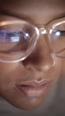 Dikey. Karanlıkta dizüstü bilgisayarı üzerinde çalışan Afrikalı Amerikalı bir hacker kadının gözlüklerindeki bilgisayar ekranının yansımasına yakın çekim. Gençler ve bilgisayar ağları ve İnternet güvenliği. 