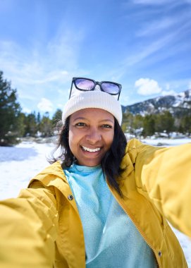 Dikey. Karlı dağlarda iki eliyle selfie çeken Afro-Amerikalı genç bir kadın. Sarı elbiseli Z nesli Latin kadın. Kış tatilinin güneşli günlerinde kameraya gülümsüyor. 